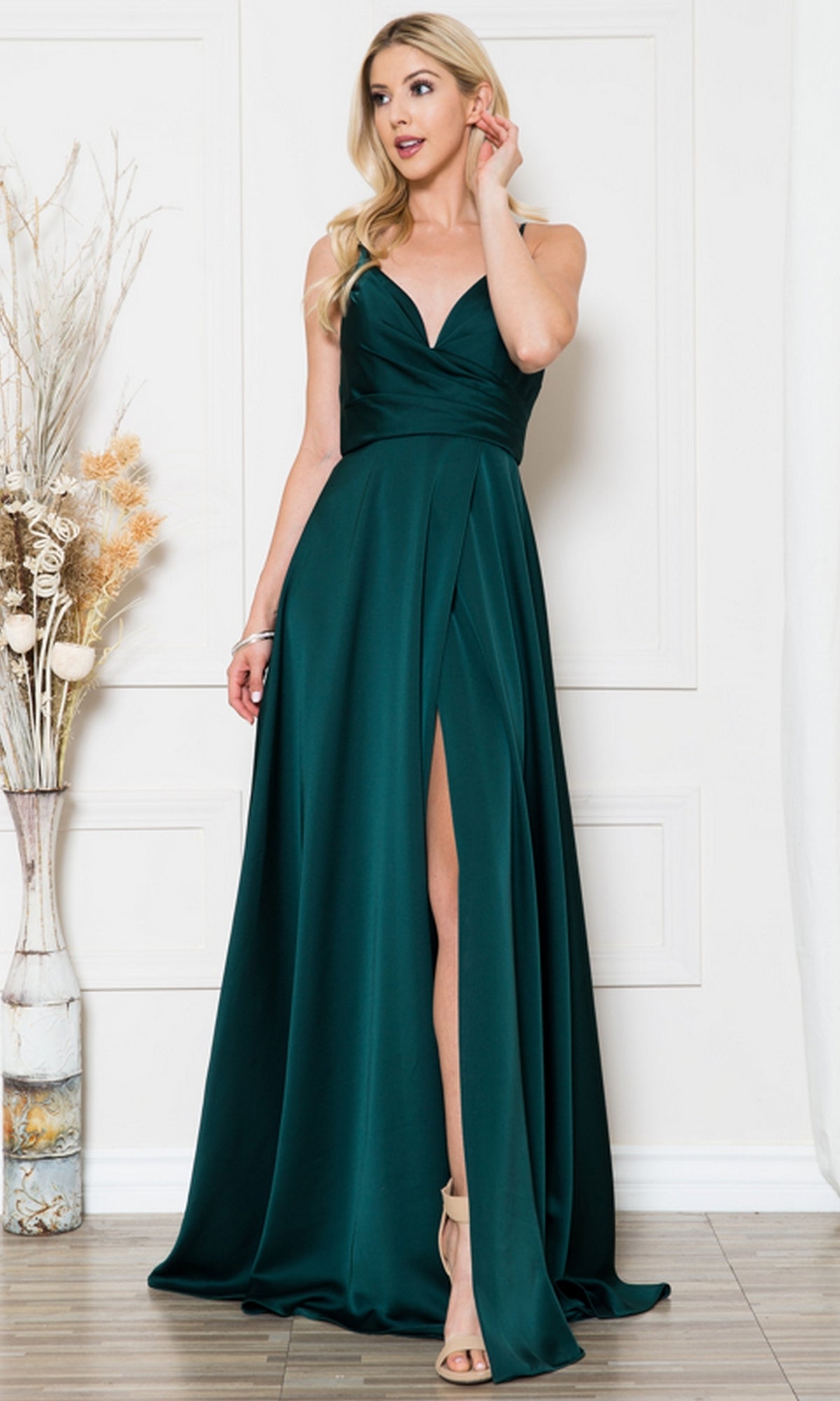 A woman stands indoors beside a vase of dried plants, wearing the Long Satin A-Line Formal Prom Dress BZ012—a sleeveless, dark green v-neck gown with a thigh-high slit.